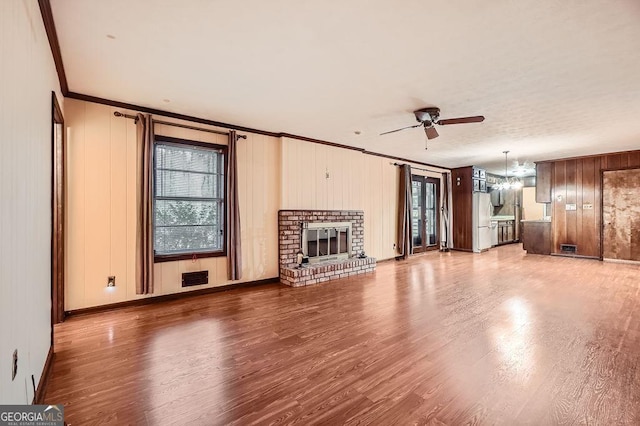 unfurnished living room featuring a brick fireplace, crown molding, wood walls, wood-type flooring, and ceiling fan with notable chandelier