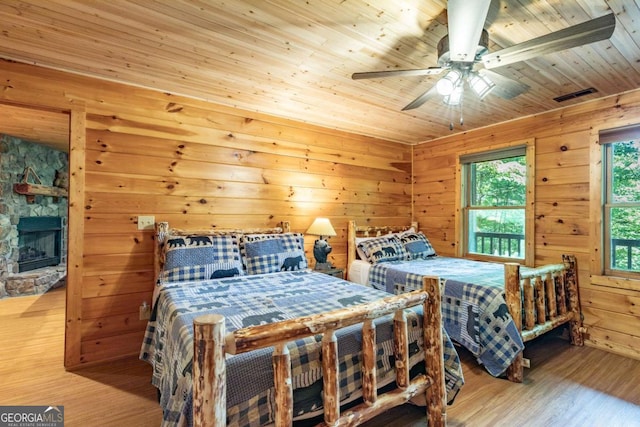 bedroom featuring wooden ceiling, a stone fireplace, wooden walls, ceiling fan, and wood-type flooring