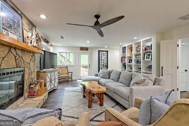 living room featuring built in shelves, ceiling fan, a stone fireplace, and wood-type flooring