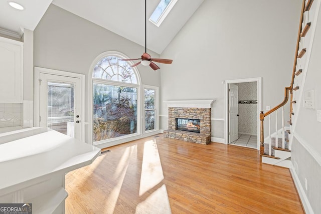 living room with a skylight, ceiling fan, a stone fireplace, high vaulted ceiling, and light hardwood / wood-style floors