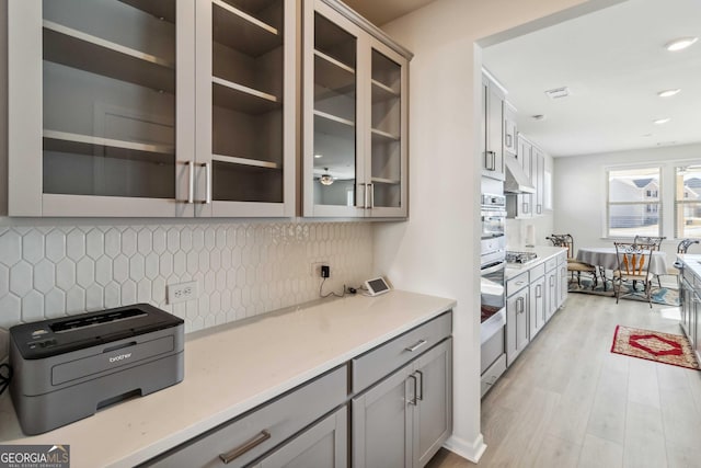 kitchen with gray cabinetry, appliances with stainless steel finishes, glass insert cabinets, light wood-style flooring, and light countertops