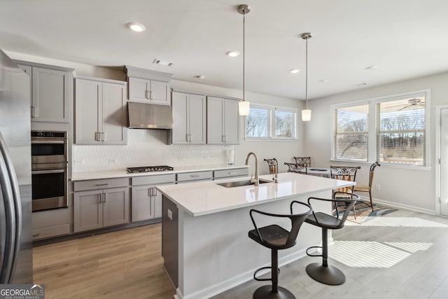 kitchen featuring sink, light hardwood / wood-style floors, pendant lighting, a kitchen island with sink, and appliances with stainless steel finishes