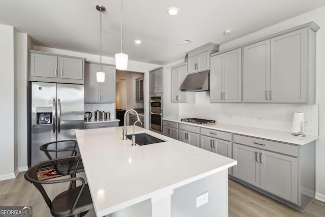 kitchen with appliances with stainless steel finishes, gray cabinetry, a sink, under cabinet range hood, and light wood-type flooring