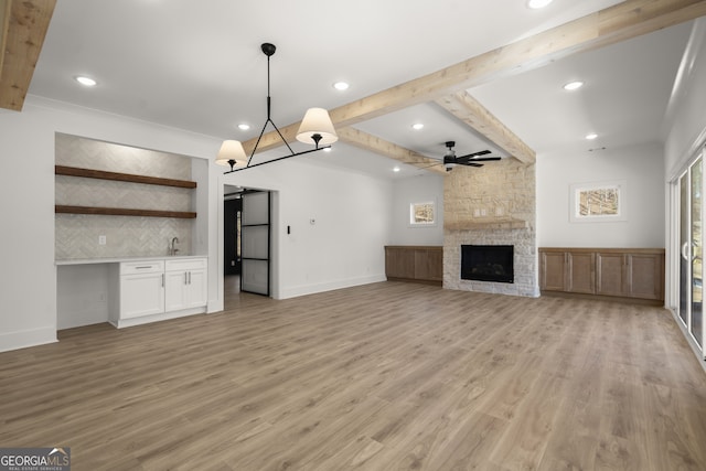 unfurnished living room featuring a fireplace, light wood-type flooring, beamed ceiling, baseboards, and ceiling fan with notable chandelier