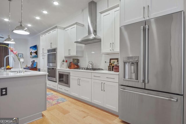 kitchen featuring wall chimney range hood, sink, white cabinetry, hanging light fixtures, and stainless steel appliances