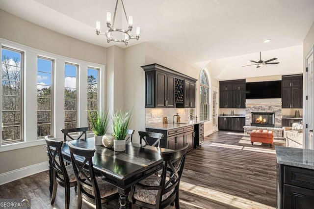 dining area with ceiling fan with notable chandelier, dark hardwood / wood-style flooring, a stone fireplace, and plenty of natural light