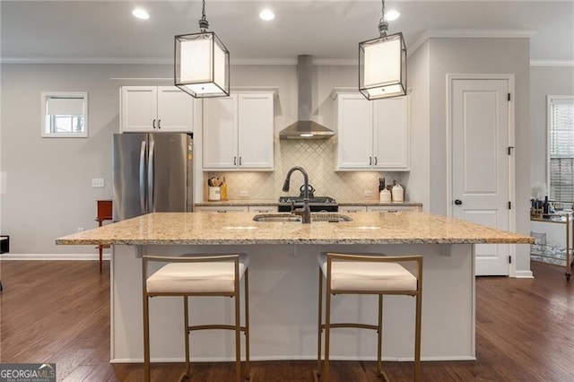 kitchen with light stone countertops, wall chimney exhaust hood, decorative light fixtures, a center island with sink, and stainless steel refrigerator