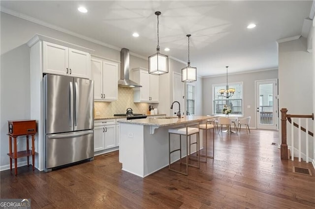 kitchen with appliances with stainless steel finishes, wall chimney exhaust hood, white cabinetry, hanging light fixtures, and an island with sink