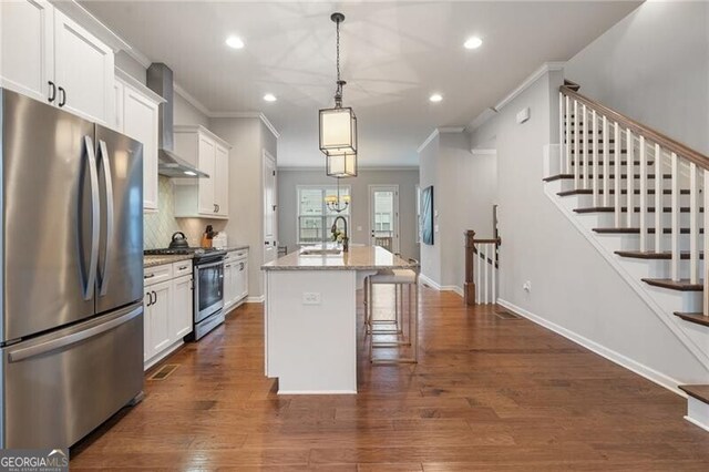 kitchen with light stone countertops, wall chimney exhaust hood, stainless steel appliances, white cabinets, and an island with sink