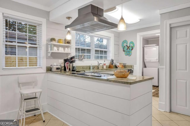 kitchen featuring kitchen peninsula, island range hood, light tile patterned floors, decorative light fixtures, and stacked washer / drying machine