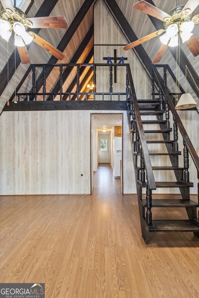 unfurnished living room featuring vaulted ceiling with beams, a ceiling fan, wooden walls, wood finished floors, and stairs