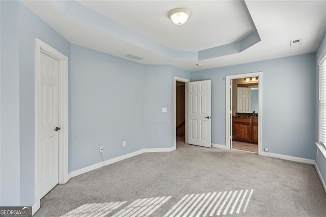 unfurnished bedroom with a raised ceiling, ensuite bath, and light colored carpet