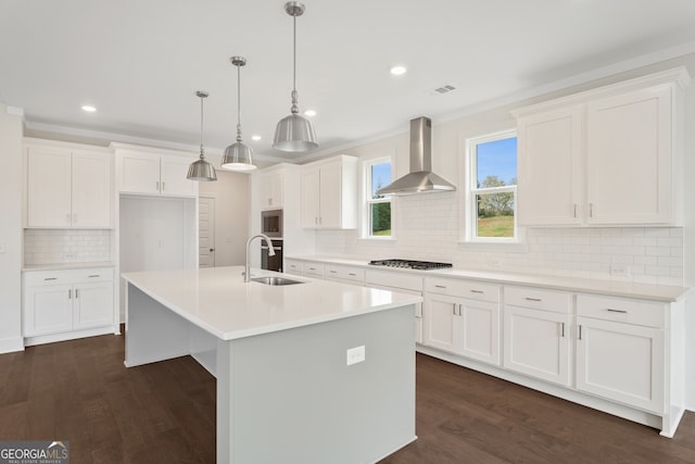 kitchen with appliances with stainless steel finishes, wall chimney range hood, pendant lighting, white cabinetry, and an island with sink