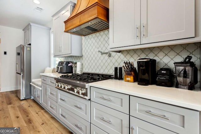 kitchen with gray cabinetry, light hardwood / wood-style floors, decorative backsplash, custom range hood, and appliances with stainless steel finishes