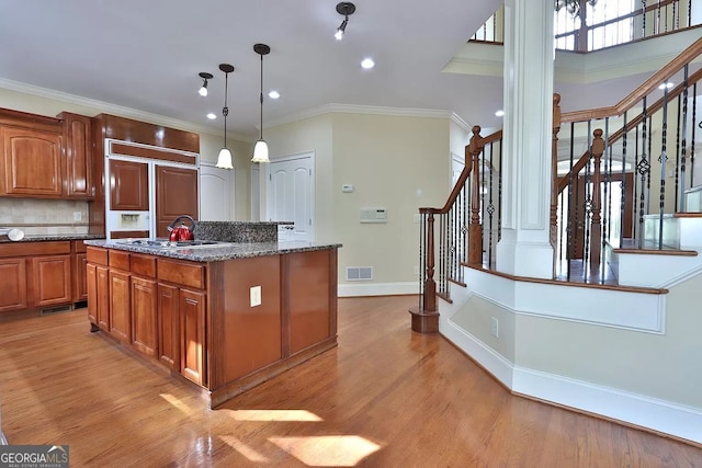 kitchen featuring hanging light fixtures, dark stone countertops, light hardwood / wood-style flooring, ornamental molding, and a kitchen island with sink