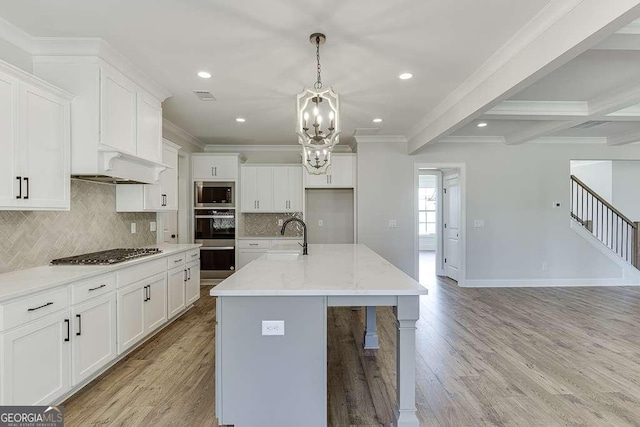 kitchen featuring ornamental molding, an island with sink, appliances with stainless steel finishes, beamed ceiling, and light stone counters