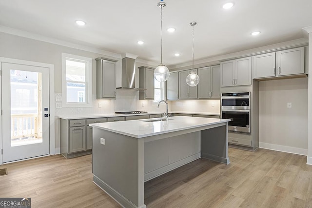 kitchen with sink, wall chimney range hood, light hardwood / wood-style flooring, double oven, and a center island with sink