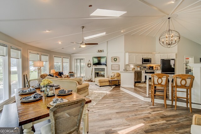 dining area with high vaulted ceiling, ceiling fan with notable chandelier, sink, a skylight, and light hardwood / wood-style floors