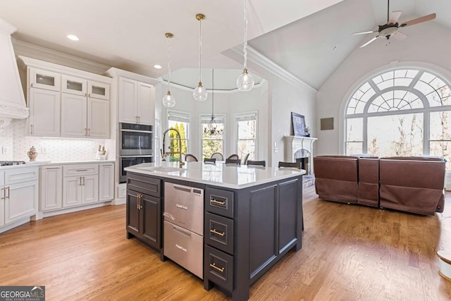 kitchen with decorative backsplash, stainless steel double oven, a kitchen island with sink, white cabinets, and hanging light fixtures