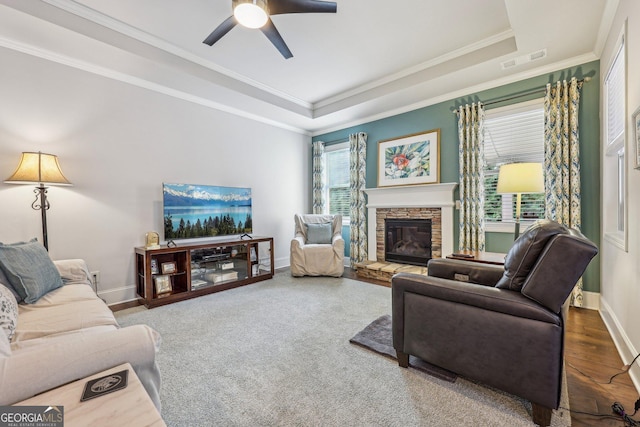 carpeted living room featuring ceiling fan, a stone fireplace, crown molding, and a tray ceiling