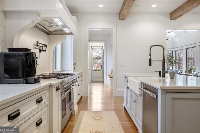 kitchen with beam ceiling, stainless steel appliances, a kitchen island with sink, white cabinets, and light wood-type flooring