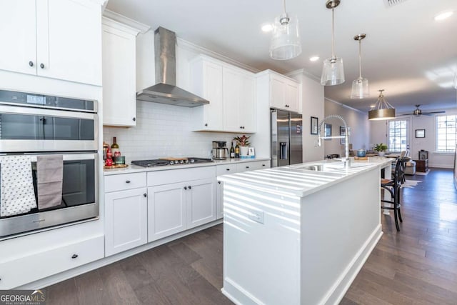 kitchen with wall chimney exhaust hood, stainless steel appliances, pendant lighting, a center island with sink, and white cabinets