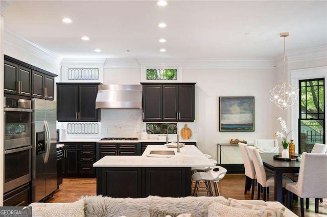 kitchen with wall chimney exhaust hood, stainless steel appliances, sink, pendant lighting, and a center island with sink