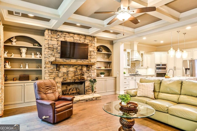 living room featuring coffered ceiling, a stone fireplace, beamed ceiling, crown molding, and light wood-type flooring