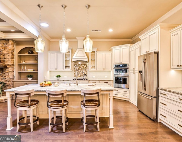 kitchen featuring appliances with stainless steel finishes, premium range hood, hanging light fixtures, a breakfast bar area, and an island with sink