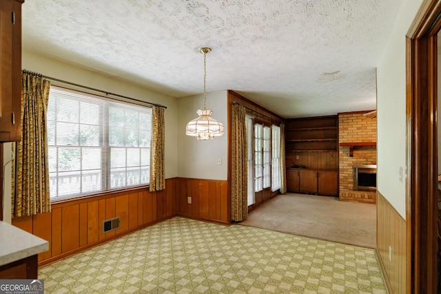 unfurnished dining area featuring light carpet, a brick fireplace, a textured ceiling, a notable chandelier, and wood walls