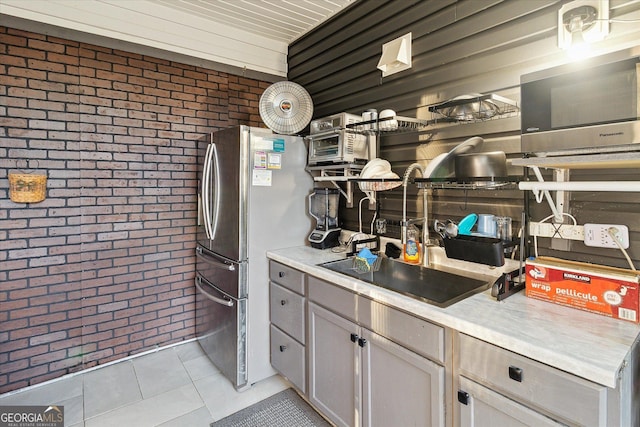 kitchen featuring gray cabinets, stainless steel refrigerator, light tile patterned floors, and brick wall