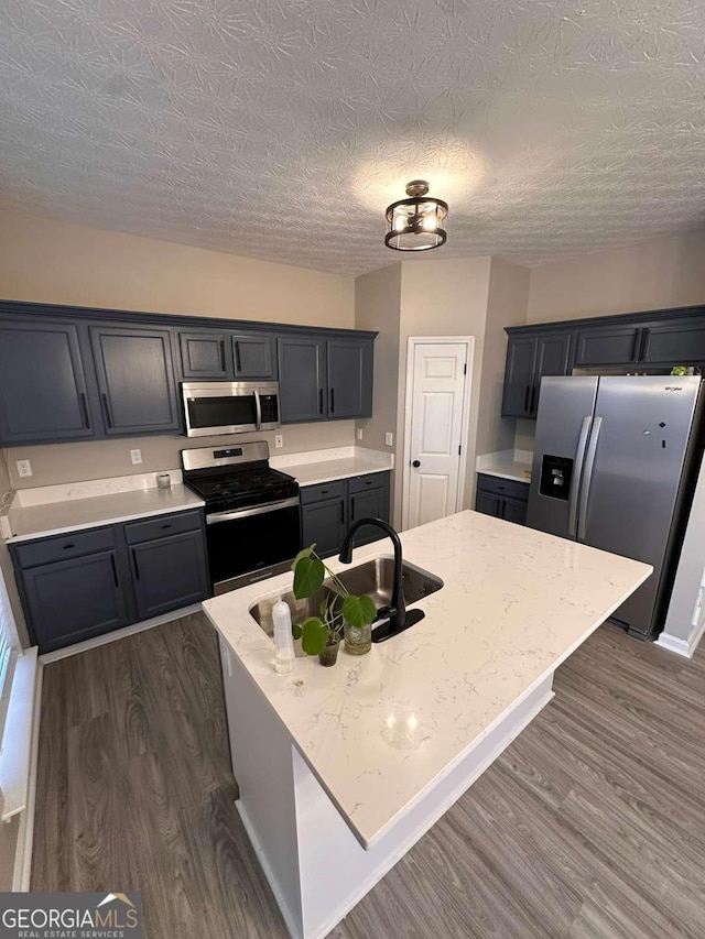 kitchen with appliances with stainless steel finishes, dark hardwood / wood-style flooring, light stone counters, a textured ceiling, and a kitchen island with sink