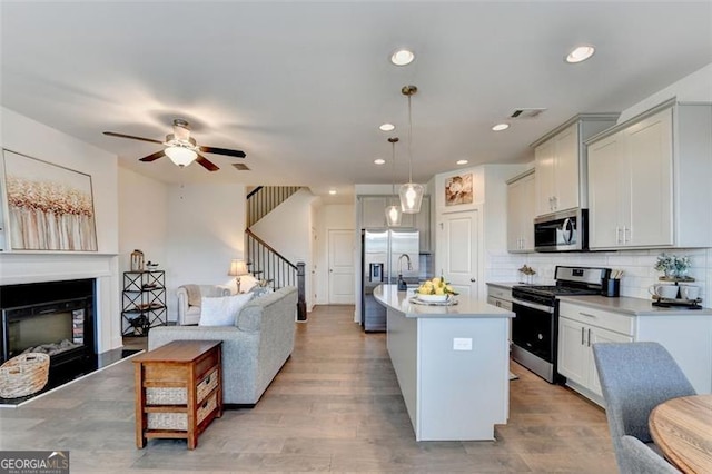 kitchen with appliances with stainless steel finishes, light wood-type flooring, tasteful backsplash, a center island with sink, and hanging light fixtures