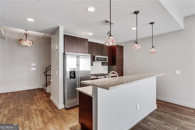 kitchen with light stone countertops, tasteful backsplash, high quality fridge, dark brown cabinets, and a kitchen island with sink