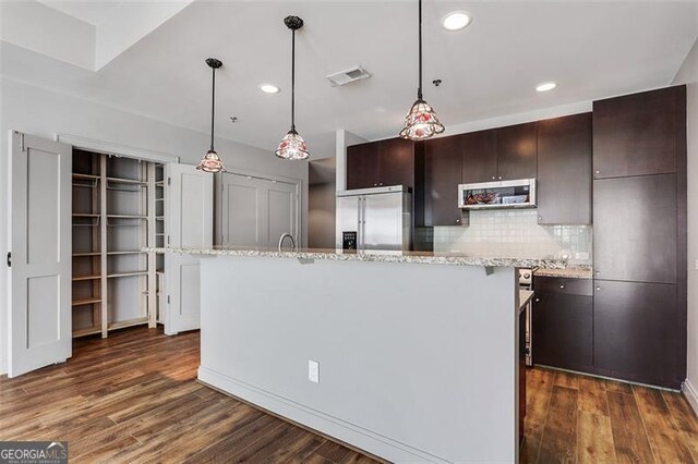 kitchen with dark brown cabinetry, dark hardwood / wood-style floors, backsplash, a center island with sink, and appliances with stainless steel finishes