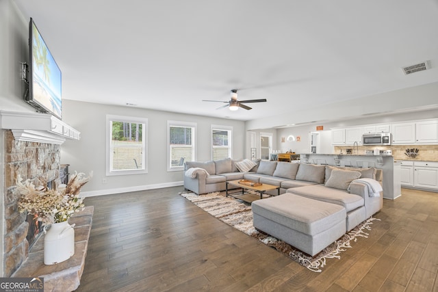 living room featuring ceiling fan, a stone fireplace, and dark wood-type flooring