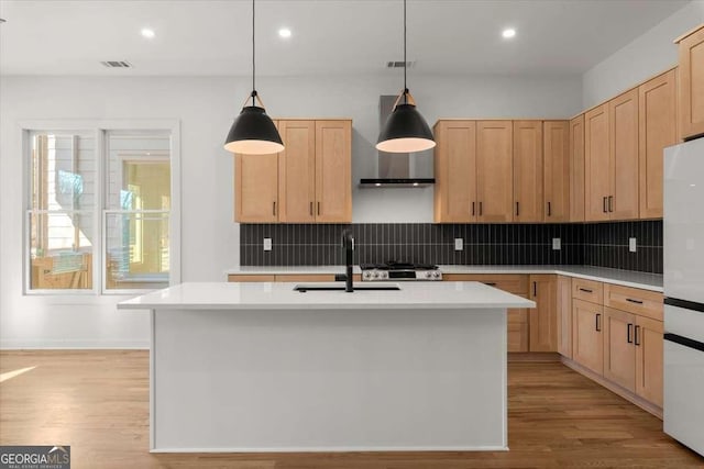 kitchen with sink, white refrigerator, an island with sink, decorative light fixtures, and light brown cabinetry