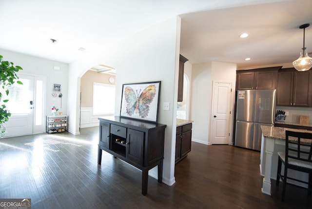 kitchen with dark wood-type flooring, hanging light fixtures, stainless steel fridge, dark brown cabinets, and light stone counters