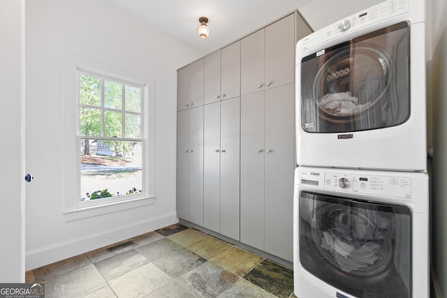 washroom featuring stone finish floor, visible vents, stacked washing maching and dryer, baseboards, and cabinet space