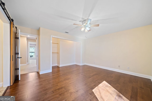 unfurnished bedroom with a barn door, ceiling fan, a closet, and dark hardwood / wood-style floors