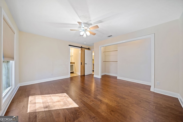 unfurnished bedroom with a barn door, ceiling fan, a closet, and dark wood-type flooring