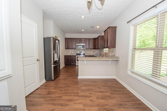 kitchen featuring dark wood-style floors, stainless steel appliances, dark brown cabinets, decorative backsplash, and a peninsula