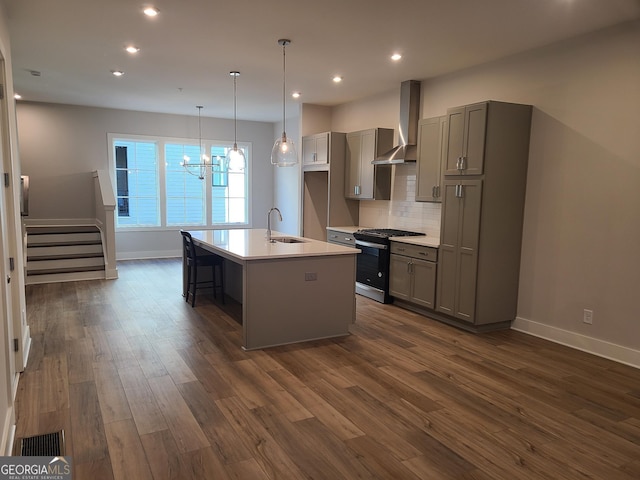 kitchen with gray cabinetry, wall chimney range hood, dark hardwood / wood-style floors, stainless steel range with gas cooktop, and an island with sink