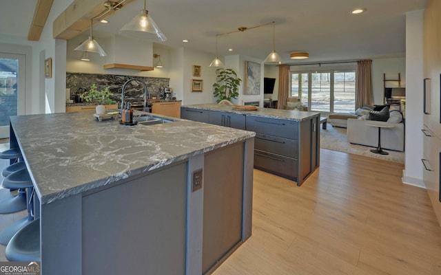 kitchen featuring sink, hanging light fixtures, backsplash, a kitchen island with sink, and light wood-type flooring