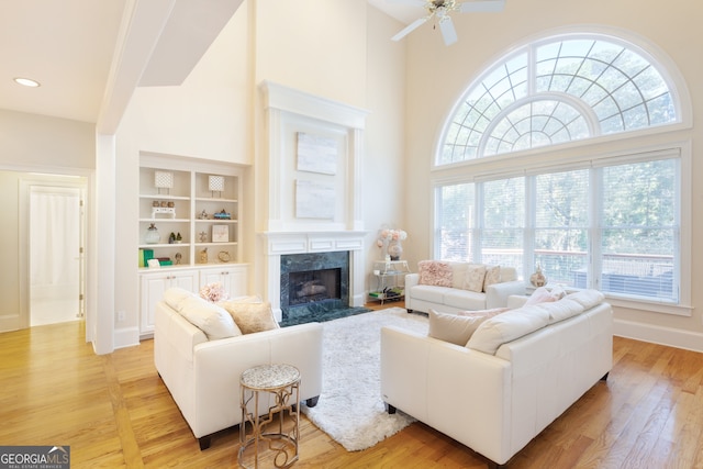 living room featuring ceiling fan, built in features, a high end fireplace, light hardwood / wood-style flooring, and a towering ceiling