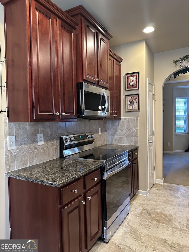 kitchen featuring stainless steel appliances, arched walkways, decorative backsplash, baseboards, and dark stone counters