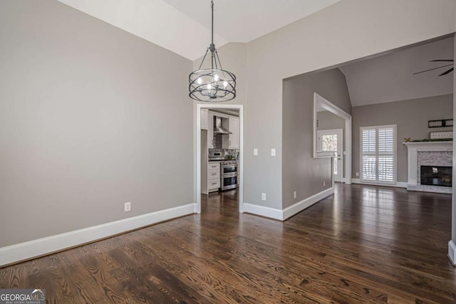 unfurnished dining area with ceiling fan with notable chandelier, vaulted ceiling, a brick fireplace, and dark wood-type flooring