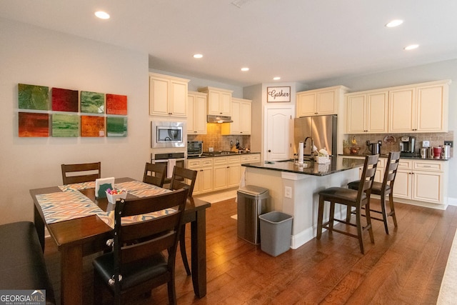 kitchen with stainless steel appliances, a kitchen breakfast bar, tasteful backsplash, an island with sink, and cream cabinetry