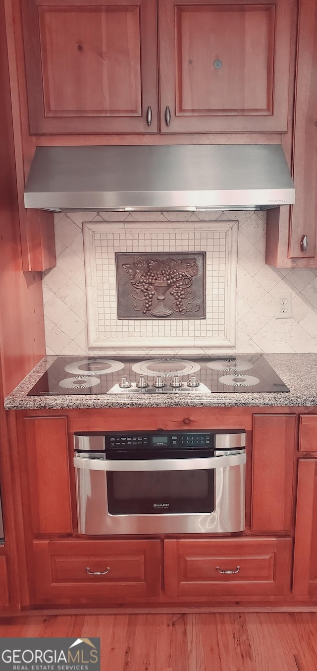 kitchen featuring tasteful backsplash, light stone counters, wall chimney exhaust hood, stainless steel oven, and black electric cooktop