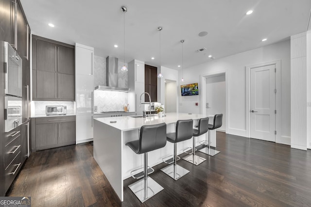 kitchen with pendant lighting, a center island with sink, a kitchen breakfast bar, wall chimney range hood, and sink